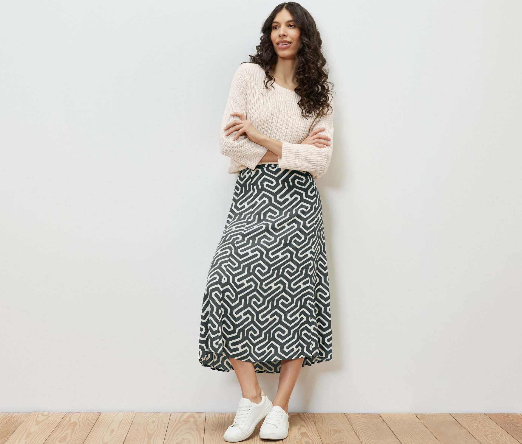 Une femme se tient debout, les bras croisés, portant une jupe midi imprimée, un pull et des baskets.