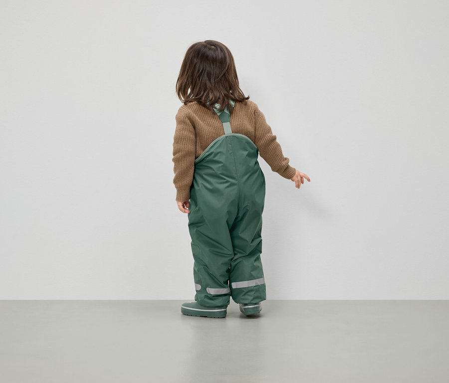 Un enfant portant un pull marron et une salopette verte se tient dos à la caméra.