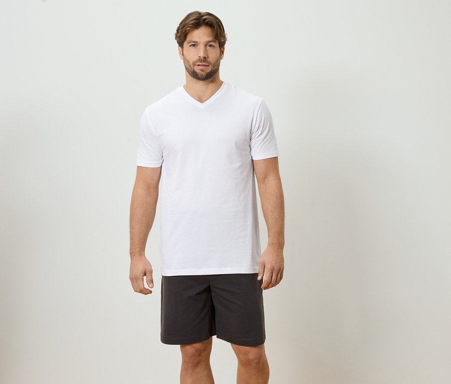 Un homme se tient devant un mur blanc, portant un t-shirt blanc et un short gris.