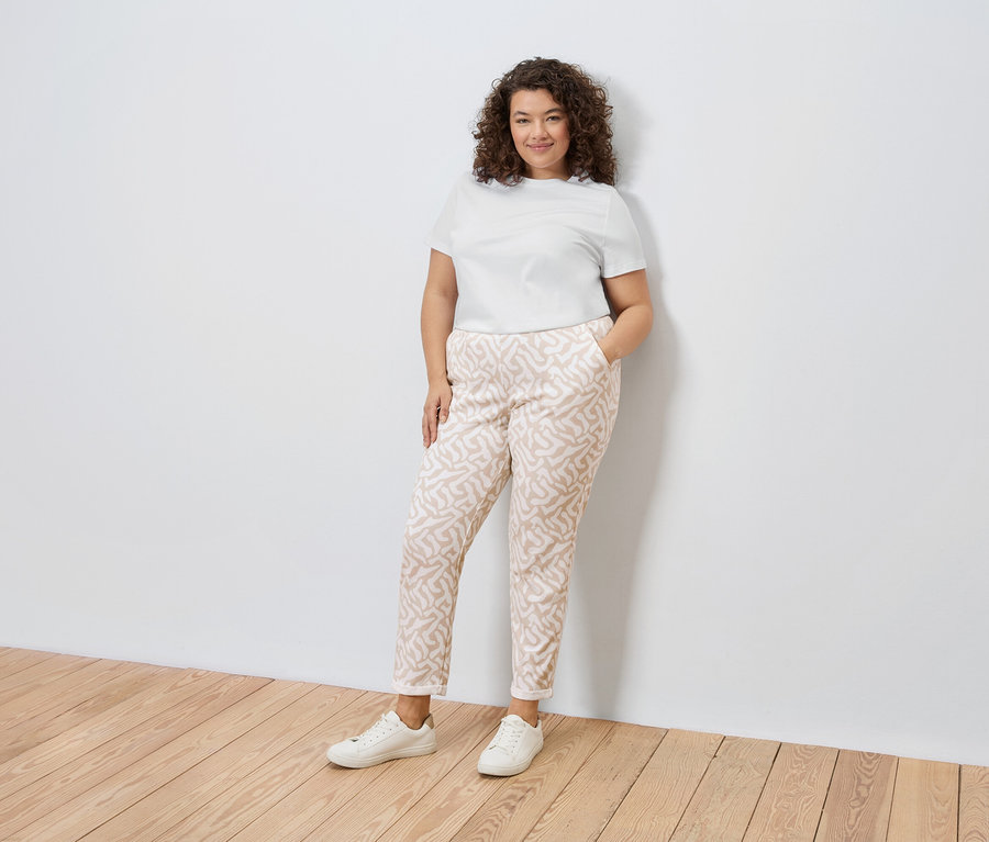 Une femme aux cheveux bouclés portant un t-shirt blanc, un pantalon à motifs et des baskets blanches, appuyée contre un mur blanc.