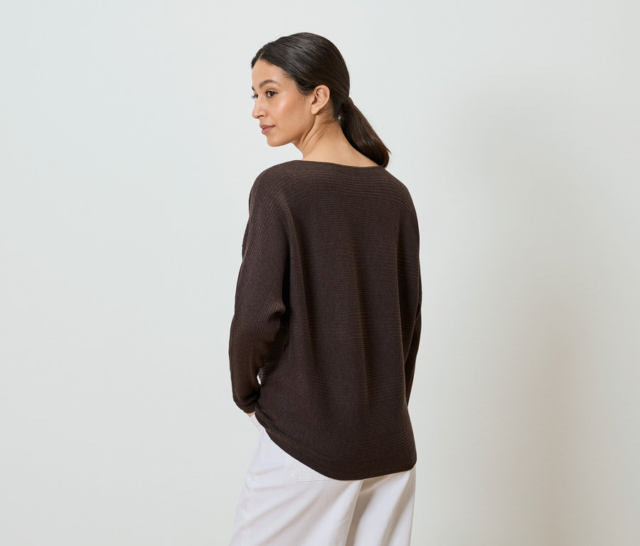 Une femme aux cheveux tressés porte un pull marron et un pantalon blanc, les mains dans les poches.