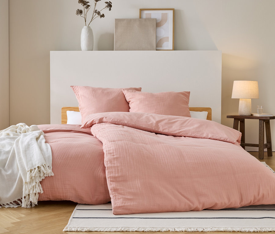 Un lit avec des draps et des oreillers roses se trouve sur un tapis rayé. Une couverture blanche est posée sur le lit. Une table de chevet se trouve à côté.