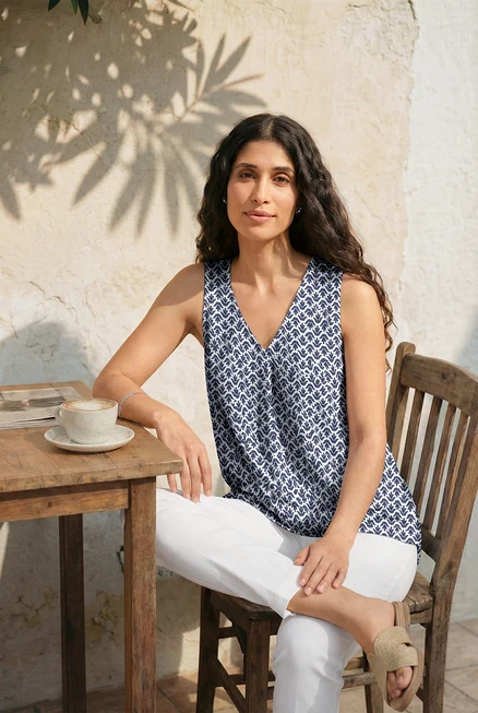 Femme assise à une table avec des ohrstecker mit glaskristallsteinen et un tennisarmband mit glaskristallsteinen. Elle porte un top mit falten-detail, minimal-alloverprint, un 7/8-stretchhose, weiß et des claquettes.