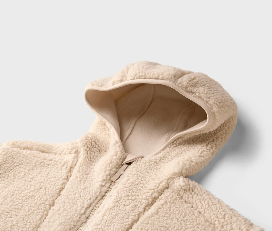 Gros plan d'une veste teddy beige à capuche et fermeture éclair sur un fond neutre.