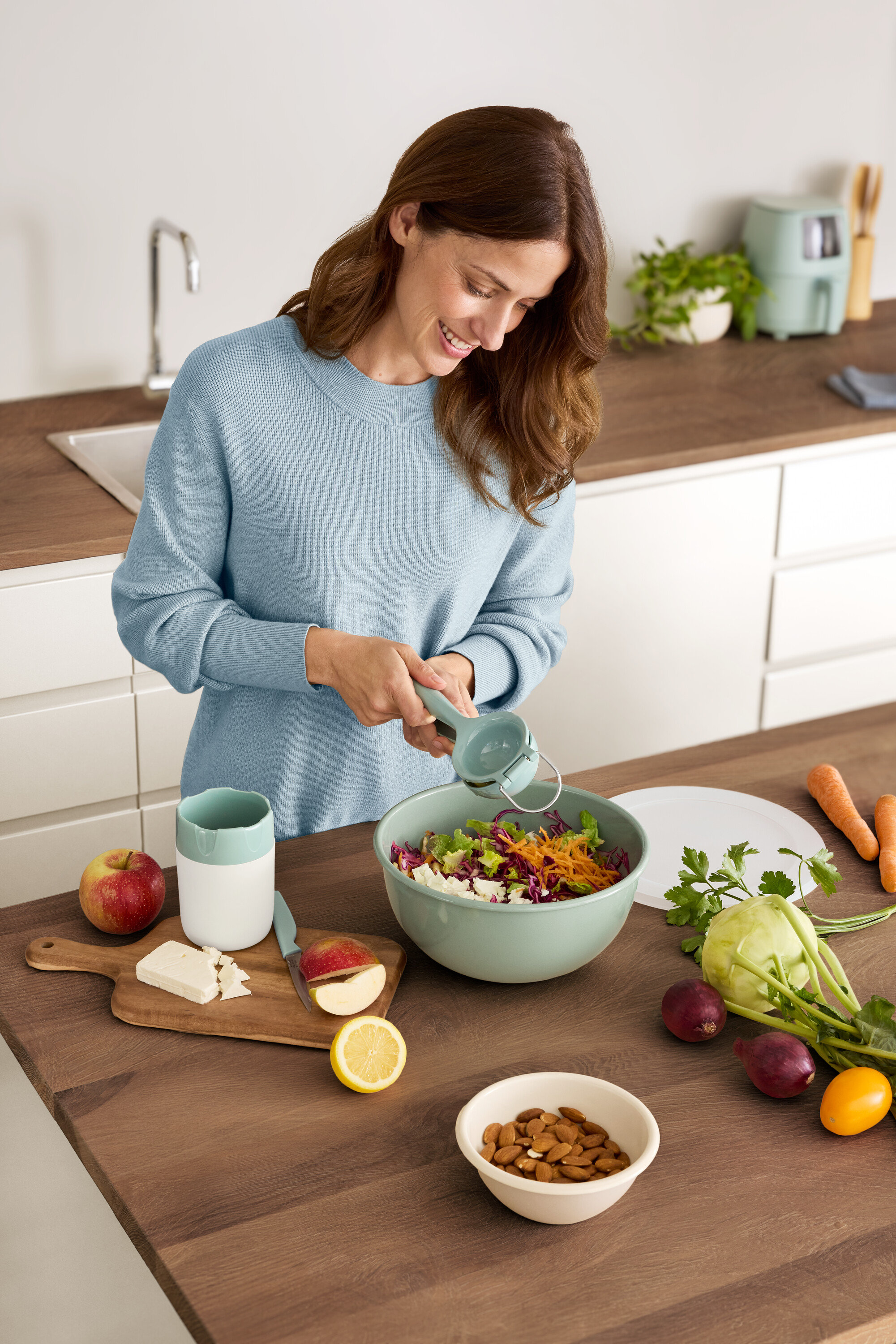 Femme pressant un citron dans un saladier. Sur la table, il y a une pomme, du fromage, des amandes et des légumes.