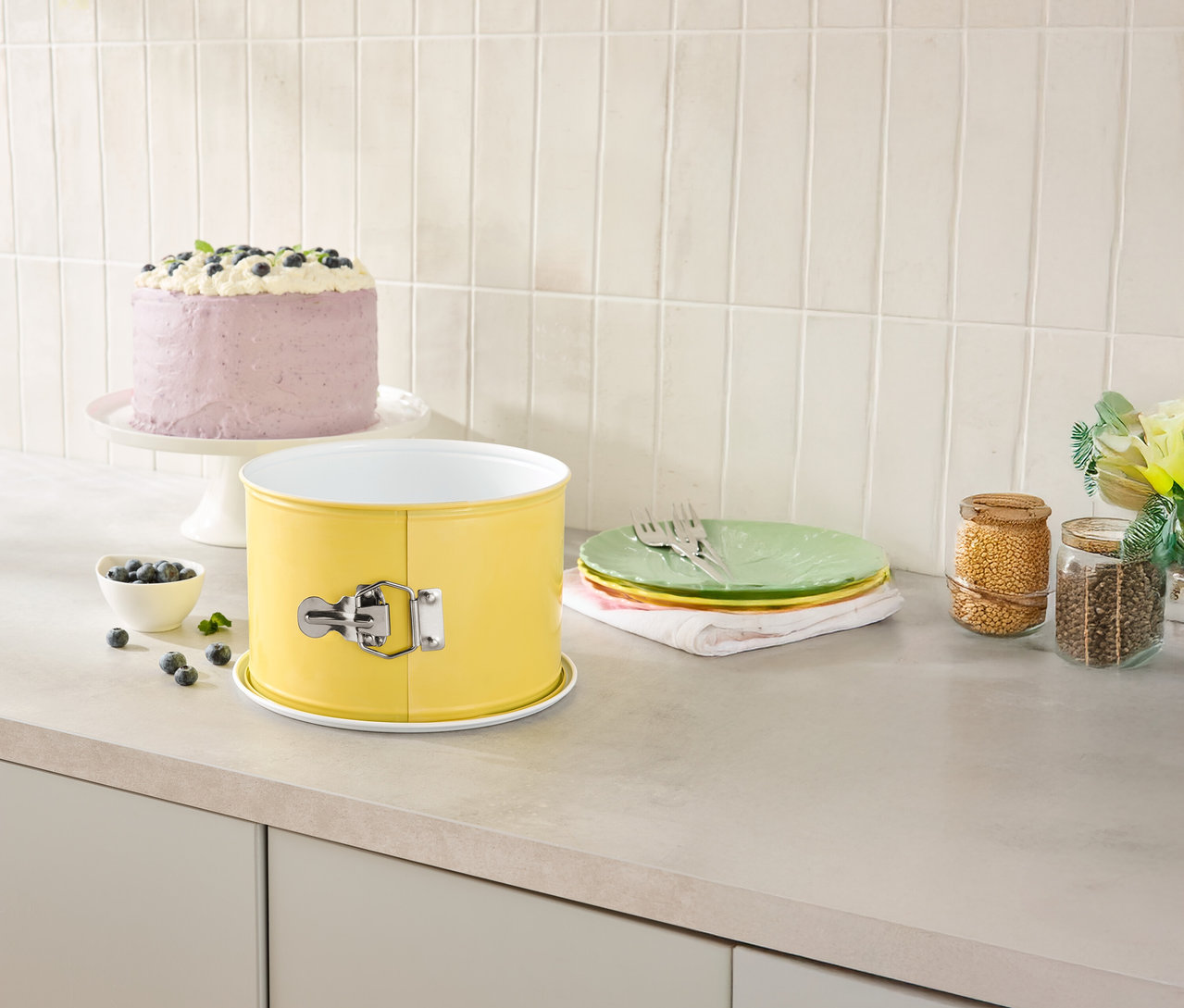 Sur un comptoir de cuisine, il y a un moule à charnière jaune, un gâteau sur un présentoir à gâteau, des assiettes et des bocaux de rangement.