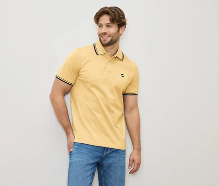 Homme portant un polo jaune et un jean, debout devant un mur clair.