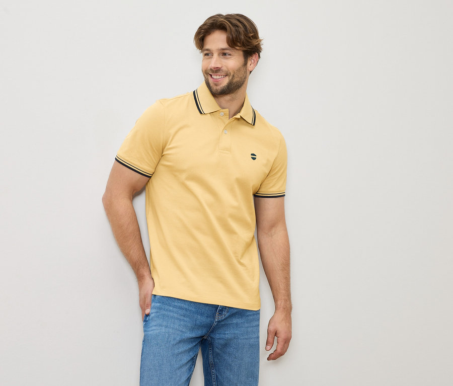 Homme portant un polo jaune et un jean, debout devant un mur clair.