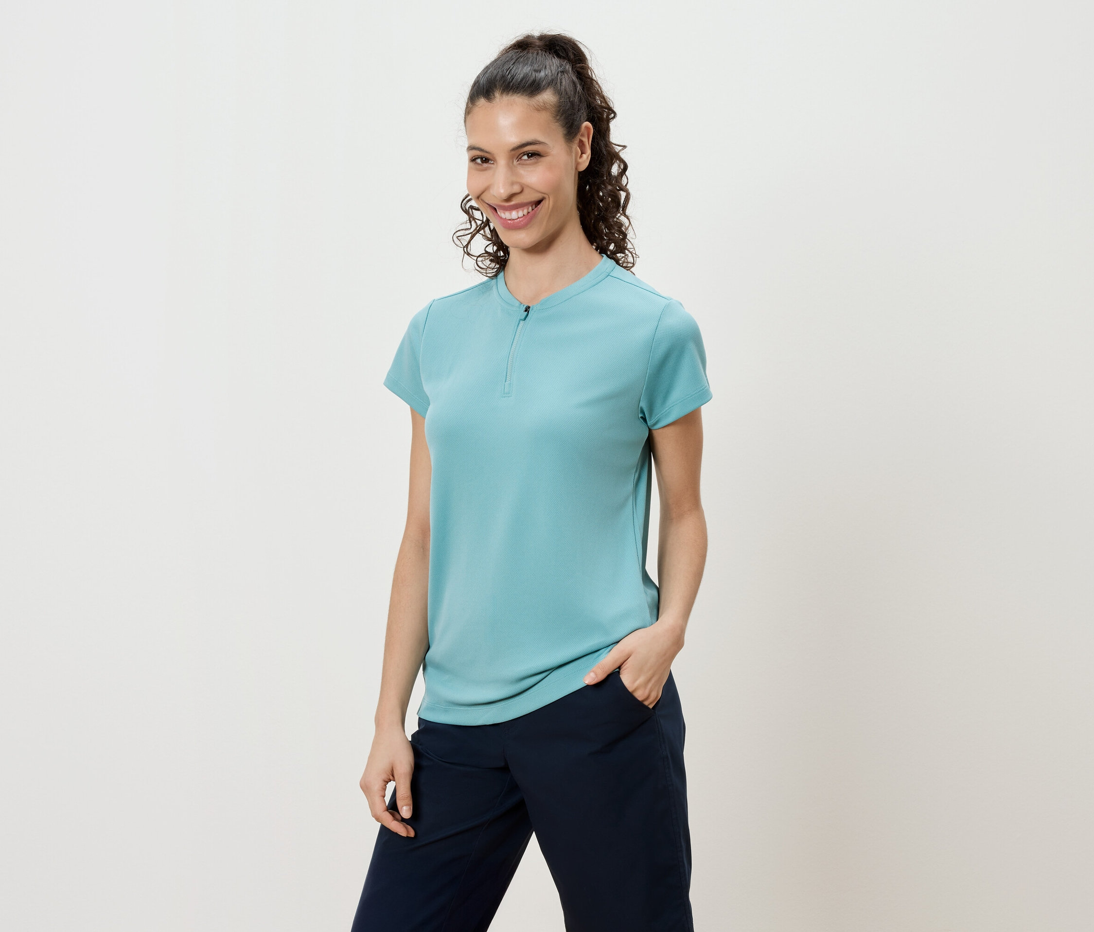 Femme aux cheveux bouclés attachés en queue de cheval portant un t-shirt turquoise et un pantalon bleu marine.