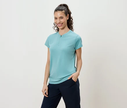 Femme aux cheveux bouclés attachés en queue de cheval portant un t-shirt turquoise et un pantalon bleu marine.