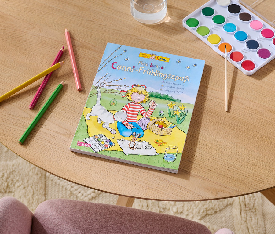 Sur une table se trouvent des crayons de couleur, un livre de coloriage intitulé "Conni-Frühlingsspaß", une boîte d'aquarelle avec un pinceau et un verre d'eau.