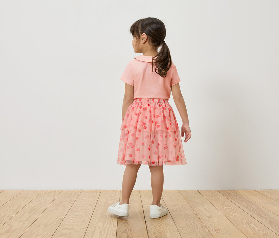 Une jeune fille se tient dos à la caméra, portant un t-shirt rose, une jupe en tulle rose avec un motif de fraises et des baskets blanches.