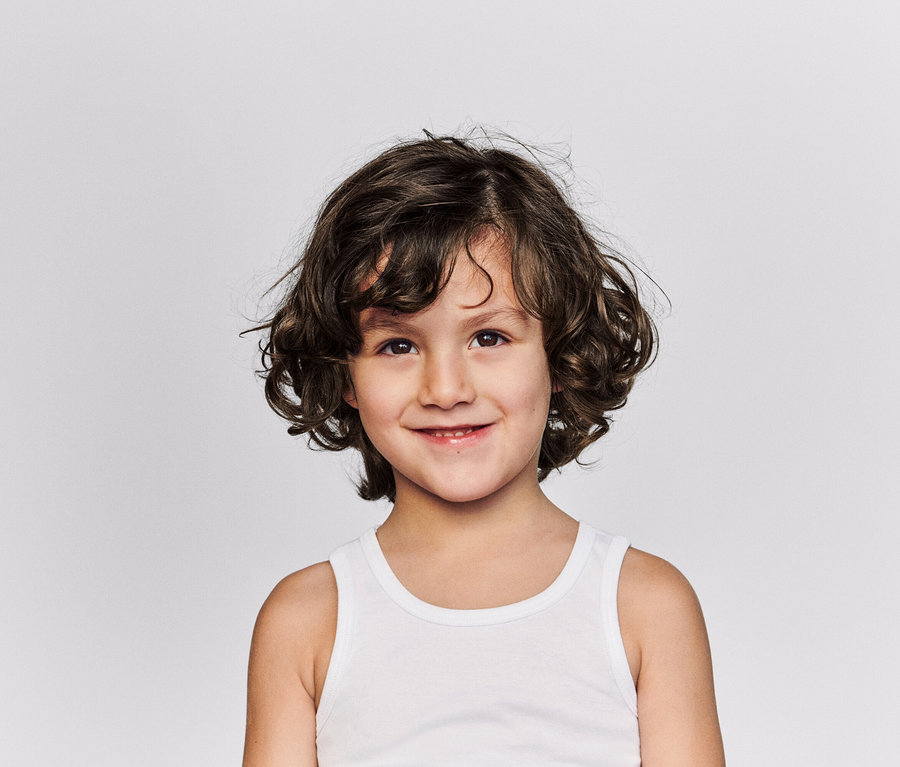 Portrait d'un enfant souriant aux cheveux bruns bouclés et portant un débardeur blanc sur un fond gris clair.