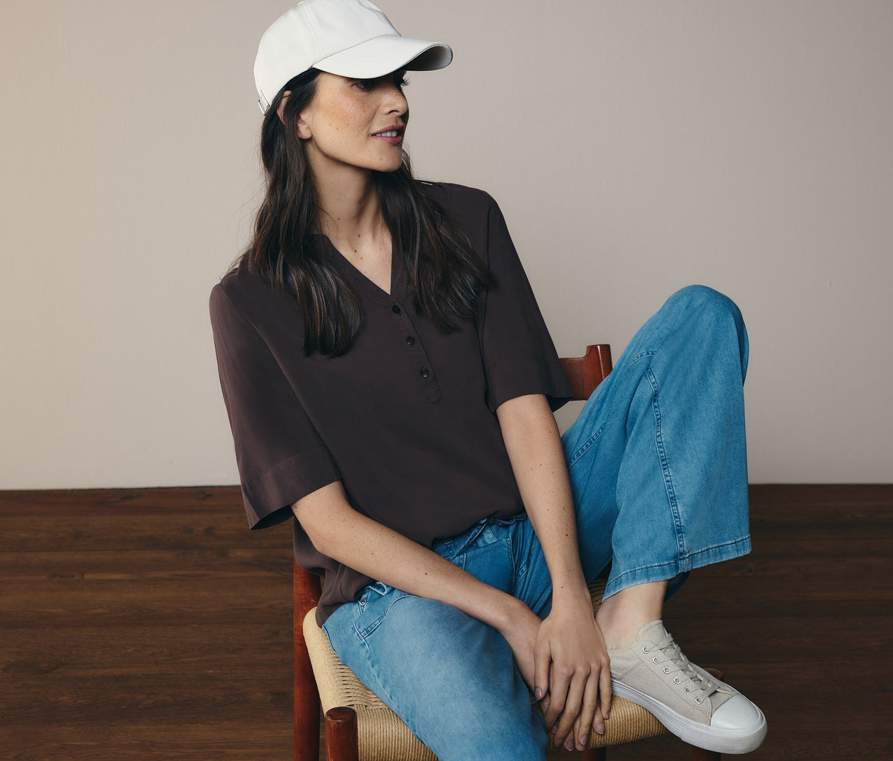 Femme assise sur une chaise, portant un jean, une casquette et un chemisier tunique CECIL marron.