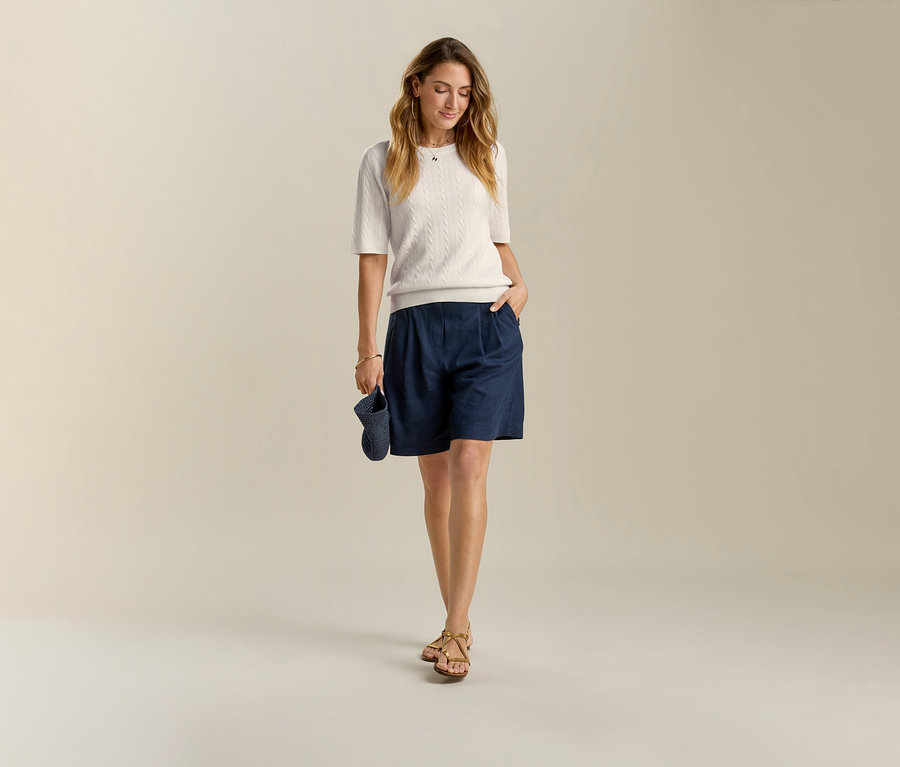 Femme portant un bermuda en lin bleu foncé et des sandales. Elle tient un chapeau bleu foncé à la main.