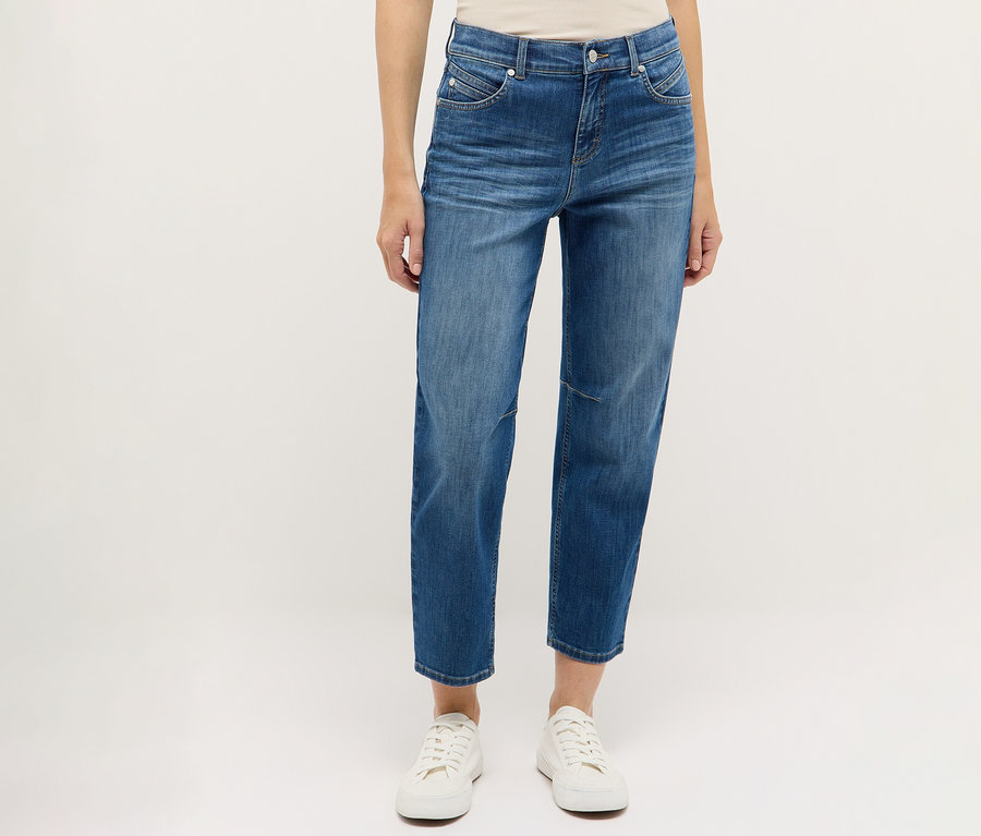 Une personne porte un jean Angels Denim « Evi » bleu et des chaussures blanches sur un fond blanc.