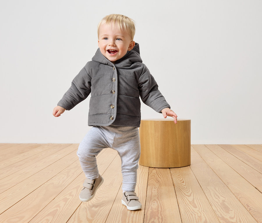 Petit garçon blond, vêtu d'une veste grise à capuche, d'un pantalon gris et de chaussures beiges, marchant sur un plancher en bois clair.