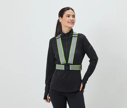 Une femme portant une queue de cheval porte un gilet de sécurité réfléchissant avec des accents vert néon sur un haut de sport noir.