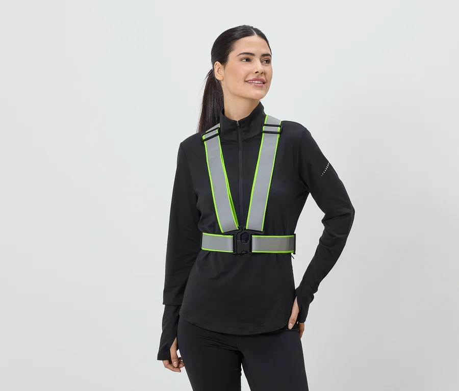 Une femme portant une queue de cheval porte un gilet de sécurité réfléchissant avec des accents vert néon sur un haut de sport noir.
