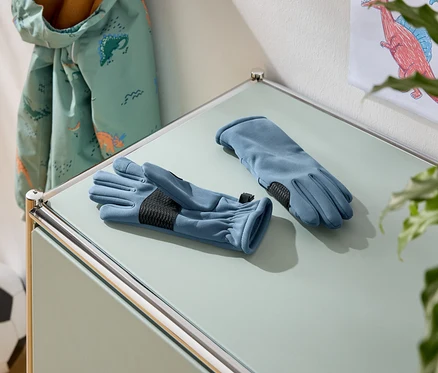 Sur une table, deux gants bleus avec des paumes noires. À l'arrière-plan, une veste à motifs de dinosaures et un ballon de football.