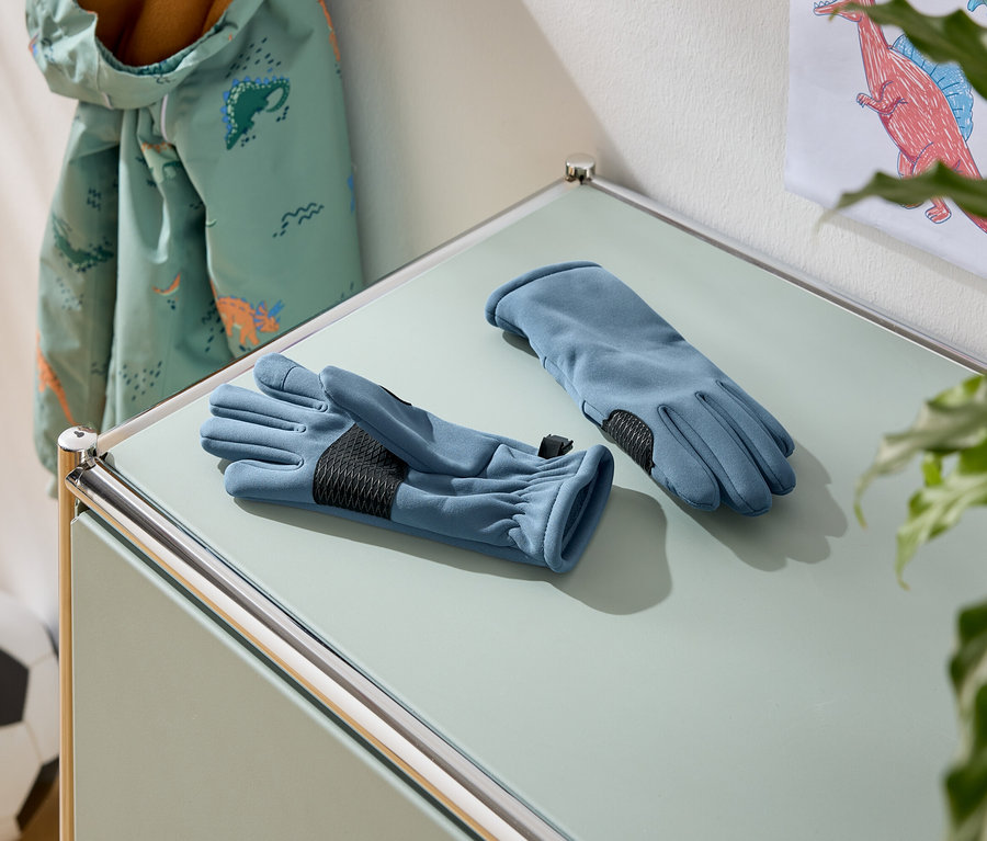 Sur une table, deux gants bleus avec des paumes noires. À l'arrière-plan, une veste à motifs de dinosaures et un ballon de football.