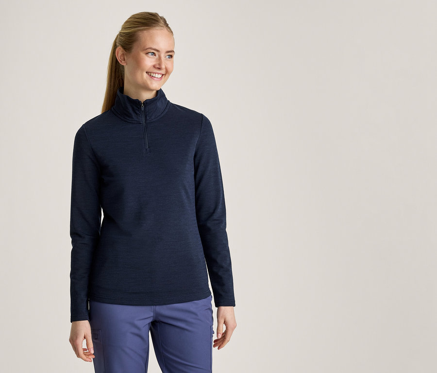 Femme souriante avec une queue de cheval, portant un haut à manches longues bleu foncé avec un demi-zip et un pantalon bleu clair.