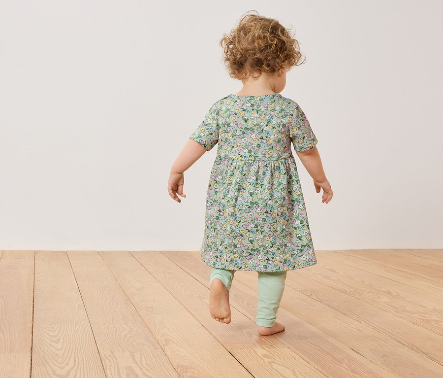 Un jeune enfant aux cheveux bouclés marche pieds nus sur un plancher en bois, portant une robe verte à fleurs et des leggings.