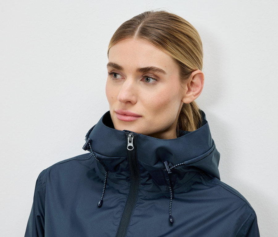 Une femme porte une veste à capuche bleu foncé sur un fond neutre.