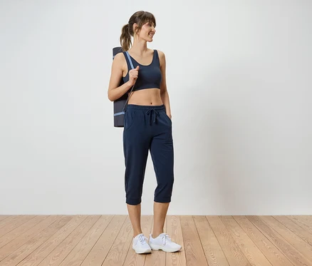 Femme portant un soutien-gorge de sport foncé, un pantacourt et un tapis de yoga sur l'épaule.