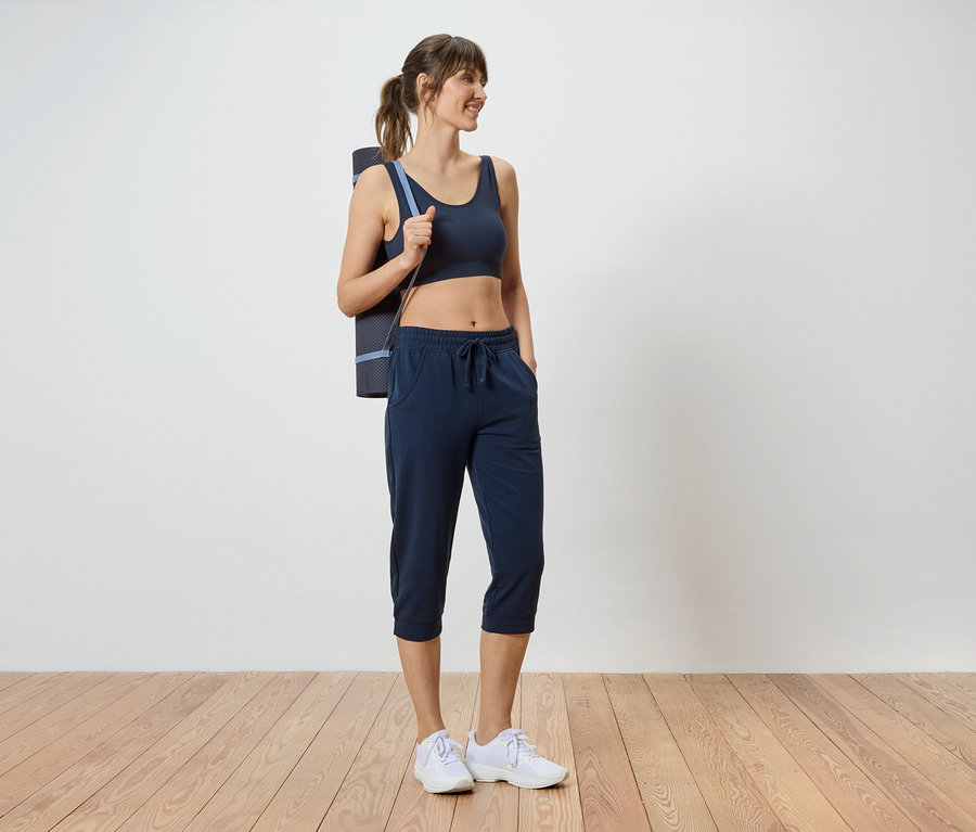 Femme portant un soutien-gorge de sport foncé, un pantacourt et un tapis de yoga sur l'épaule.
