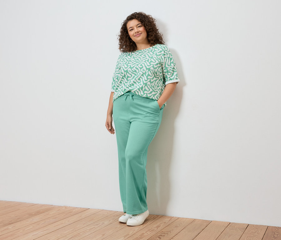Une femme pose contre un mur, portant un haut à motifs vert clair, un pantalon vert clair et des baskets blanches.