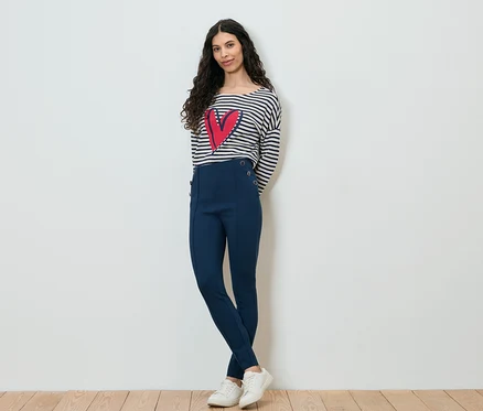 Une femme aux cheveux bruns bouclés, portant un haut rayé à manches longues avec un cœur rouge, un pantalon bleu marine et des baskets blanches, pose devant un mur blanc.