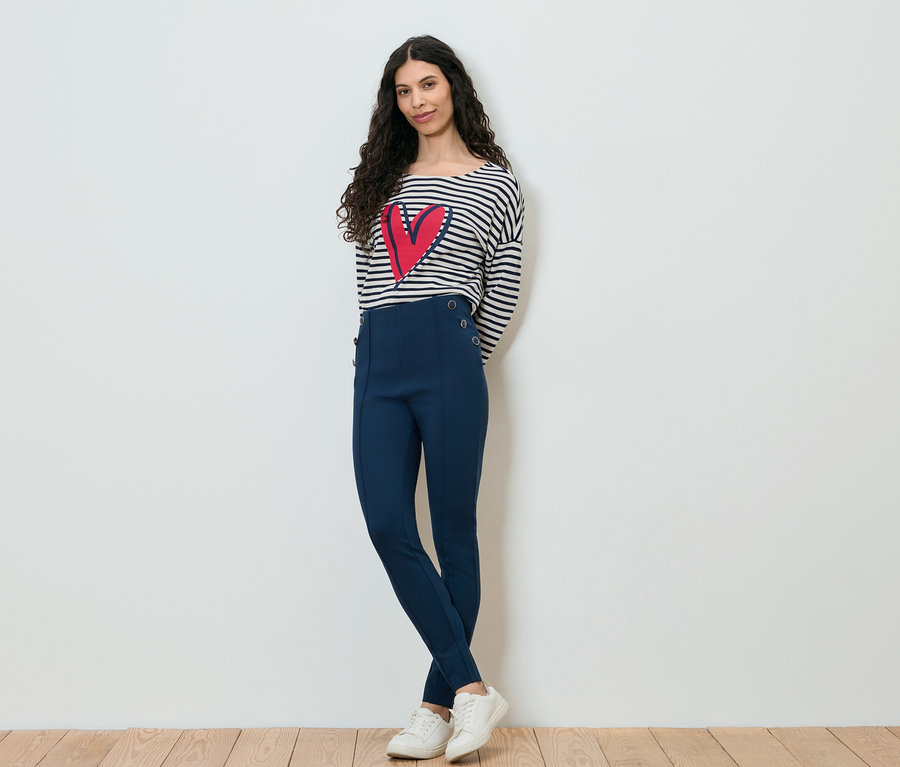 Une femme aux cheveux bruns bouclés, portant un haut rayé à manches longues avec un cœur rouge, un pantalon bleu marine et des baskets blanches, pose devant un mur blanc.