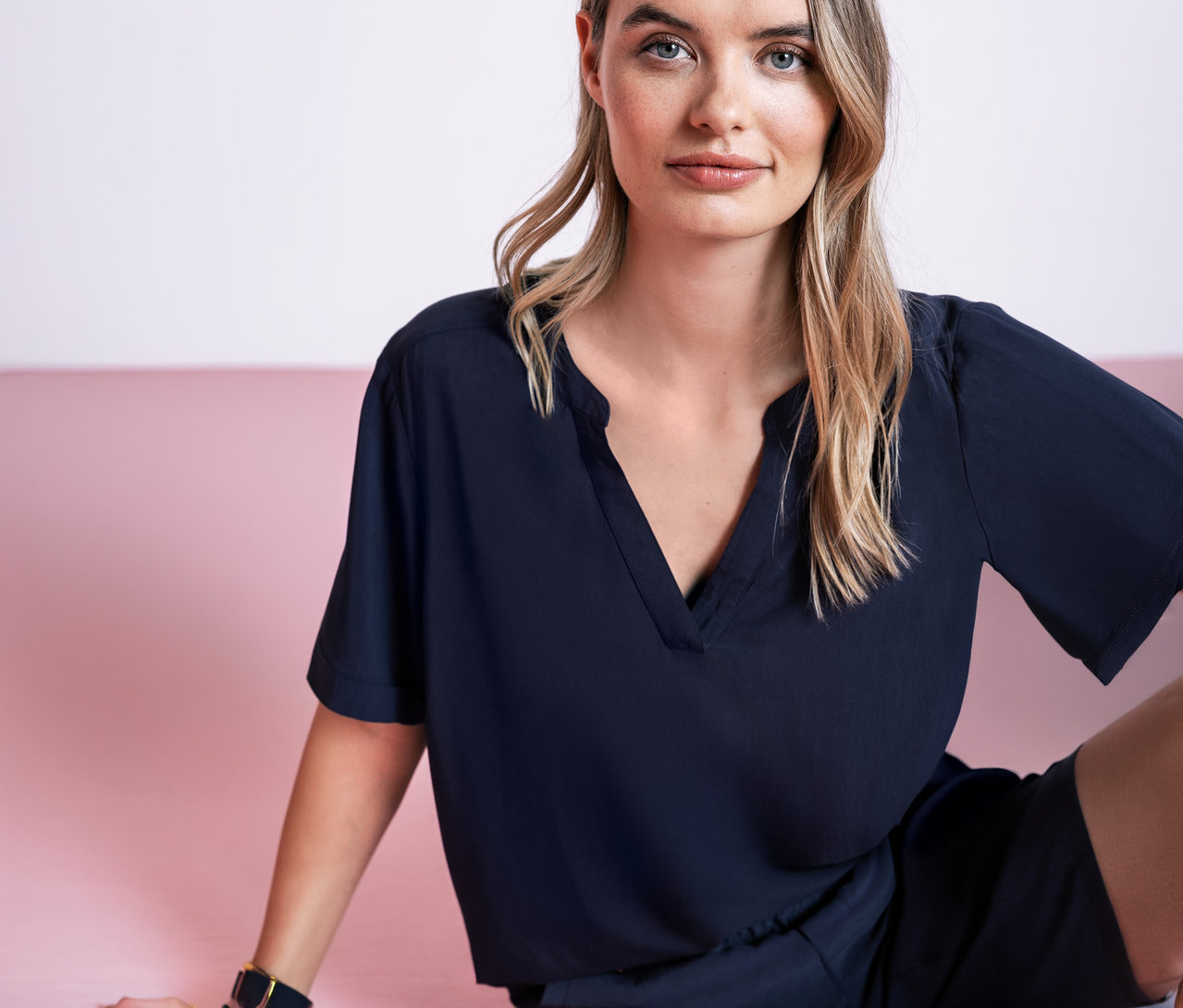 Une femme est assise devant un fond rose et porte une blouse en maille Street One bleu foncé.