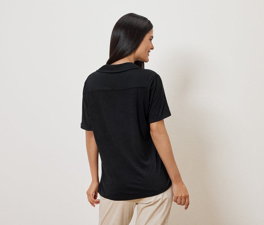 Femme vue de dos portant un polo noir et un pantalon beige sur fond blanc.