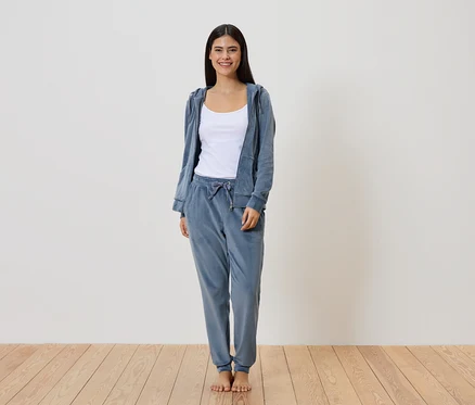 Femme en survêtement en velours bleu avec veste à capuche et débardeur blanc, pieds nus sur un plancher en bois devant un mur blanc.