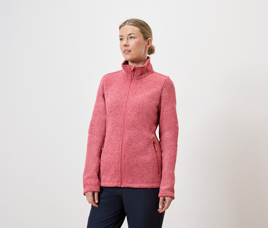 Une femme portant un chignon porte une veste en polaire rose zippée et un pantalon bleu marine sur fond blanc.