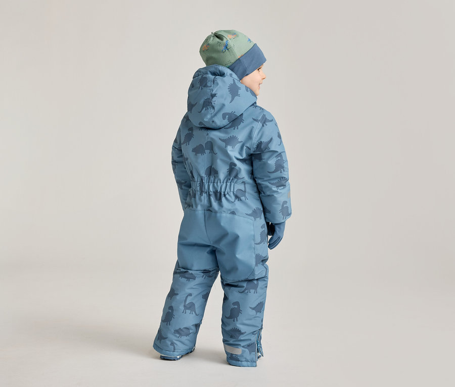 Un enfant, dos à la caméra, porte une combinaison de ski bleue à motifs de dinosaures et un bonnet vert à dinosaures.