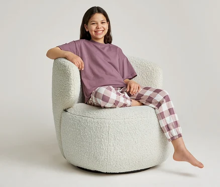 Une jeune fille souriante est assise pieds nus dans un fauteuil blanc, portant un t-shirt violet et un pantalon à carreaux.