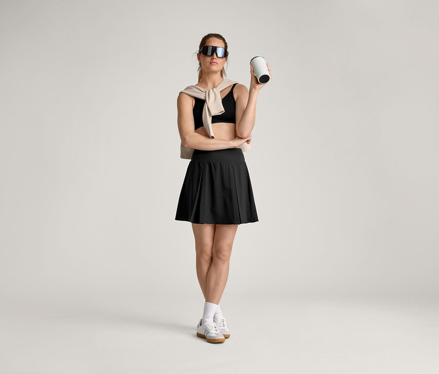 Une femme portant des lunettes de soleil, une brassière de sport, une jupe, des chaussettes et des baskets tient une tasse, avec un pull sur les épaules.