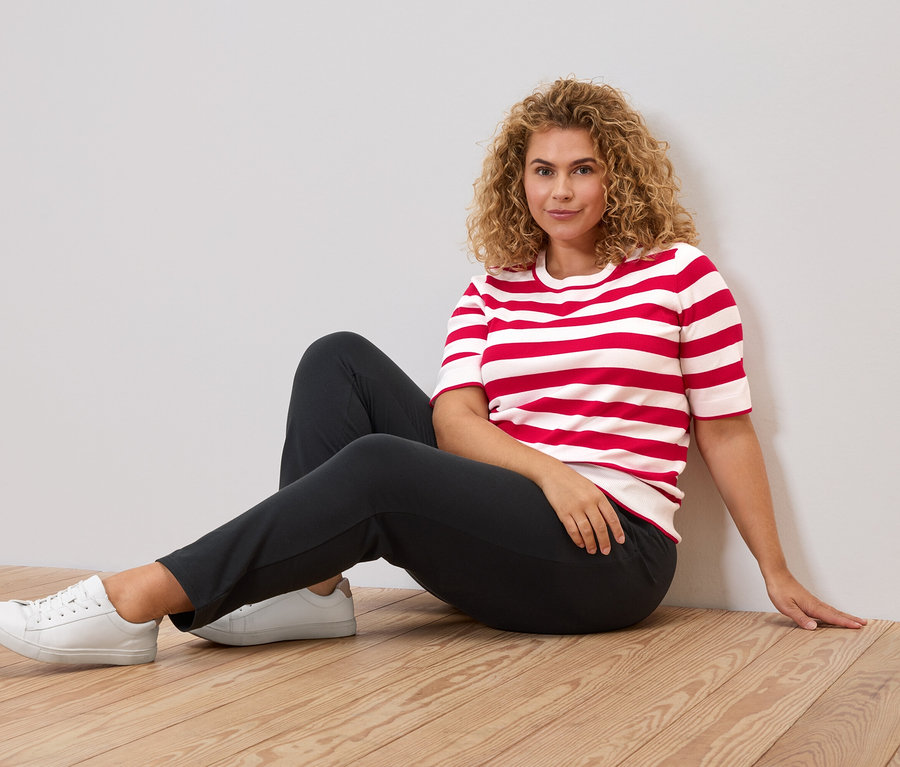 Femme aux cheveux bouclés assise sur un plancher en bois, portant un t-shirt rayé rouge et blanc, un pantalon foncé et des baskets blanches.