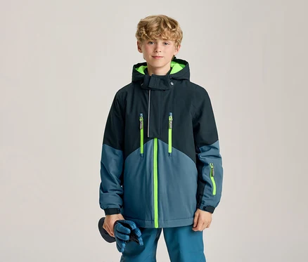Jeune garçon debout, portant une veste d'hiver bleu foncé et bleu clair avec des fermetures éclair et une doublure de capuche vert fluo, tenant des gants bleus.
