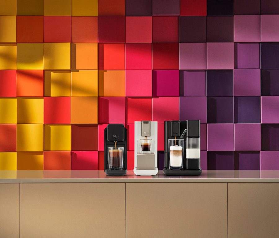 Trois machines à café Qbo sur un comptoir devant un mur carrelé coloré. Une machine à capsules Qbo ONE noire prépare du café, une Qbo ICONIC or clair prépare du café et une Qbo MILK MASTER noire avec mousseur prépare une boisson en couches.