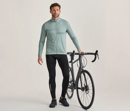 Un homme se tient à côté d'un vélo de route gris foncé, tenant son guidon. Il porte un maillot de cyclisme vert clair à manches longues avec un motif sur la poitrine, un cuissard de cyclisme noir avec une bande réfléchissante et des chaussures de cyclisme noires.
