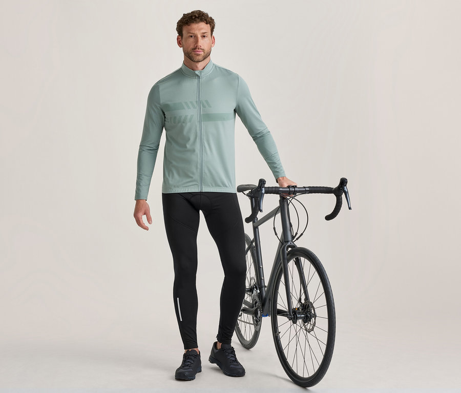 Un homme se tient à côté d'un vélo de route gris foncé, tenant son guidon. Il porte un maillot de cyclisme vert clair à manches longues avec un motif sur la poitrine, un cuissard de cyclisme noir avec une bande réfléchissante et des chaussures de cyclisme noires.