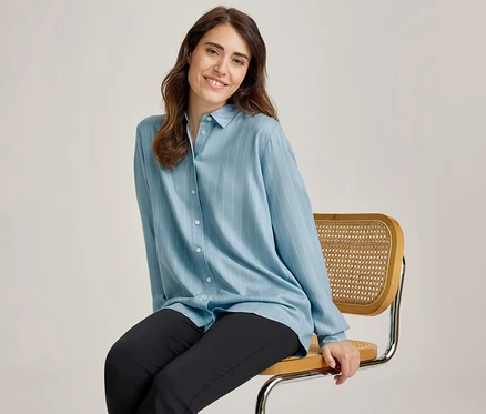 Une femme souriante aux longs cheveux bruns est assise à l'envers sur une chaise en bois et cannage. Elle porte une blouse rayée bleu clair et un pantalon noir.