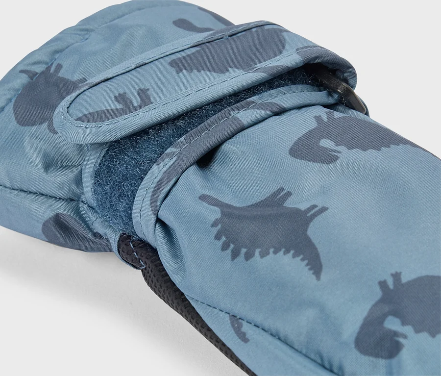 Gros plan d'une moufle bleue avec un motif de dinosaures bleu foncé. Une sangle velcro est partiellement ouverte au poignet, révélant le dessous duveteux. Une boucle noire est visible près du poignet.