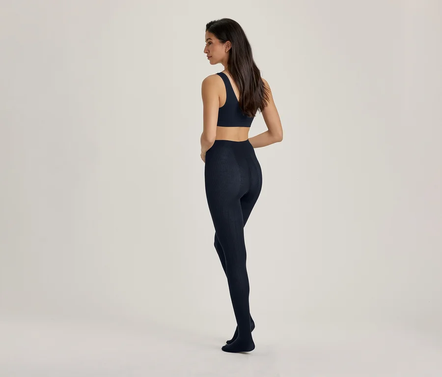Une femme est debout, de dos, légèrement tournée vers la gauche, portant un soutien-gorge de sport bleu foncé et un legging bleu foncé assorti.