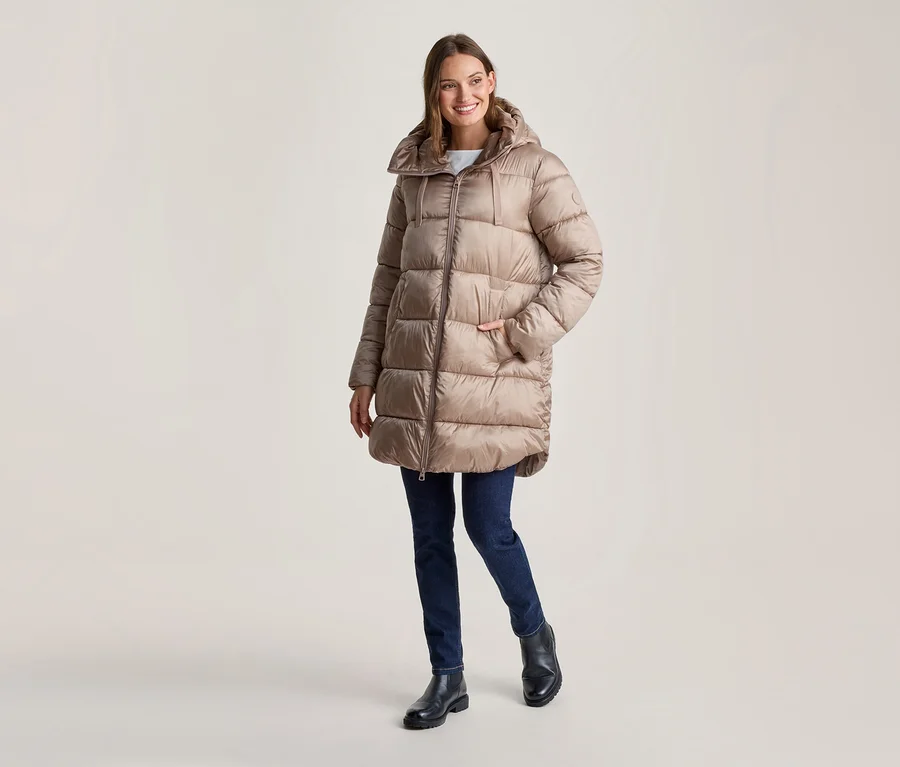 Femme souriante portant un long manteau doudoune beige à capuche, un jean bleu foncé et des bottines noires, une main dans la poche.