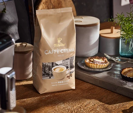 Sur une table en bois, un paquet de Caffè Crema Mild, une machine à café et des assiettes avec des gâteaux.